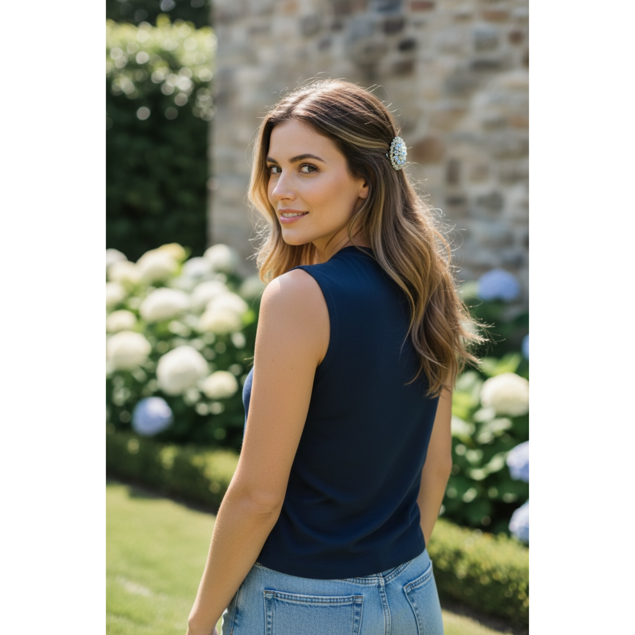 Woman in a navy sleeveless top and jeans standing outdoors with flowers and a stone wall in the background. The highlight of the image is her decorative multicolor hairclip