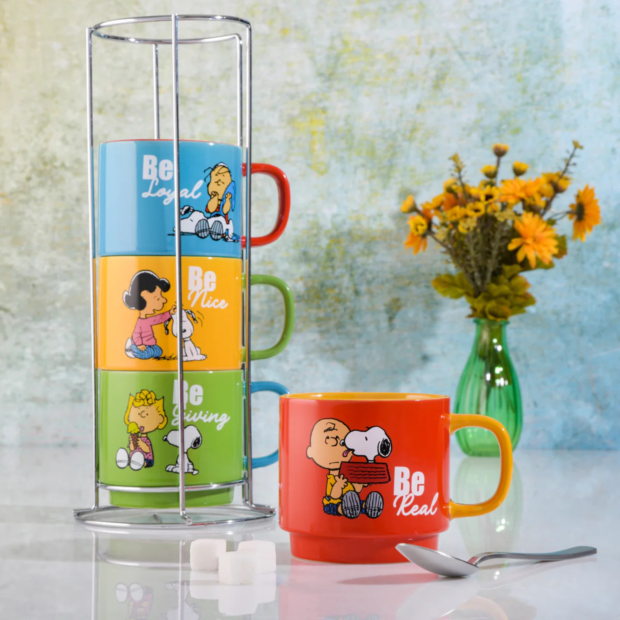 Set of colorful mugs with cartoon characters and motivational phrases, displayed with a transparent stand and a vase of flowers in the background.