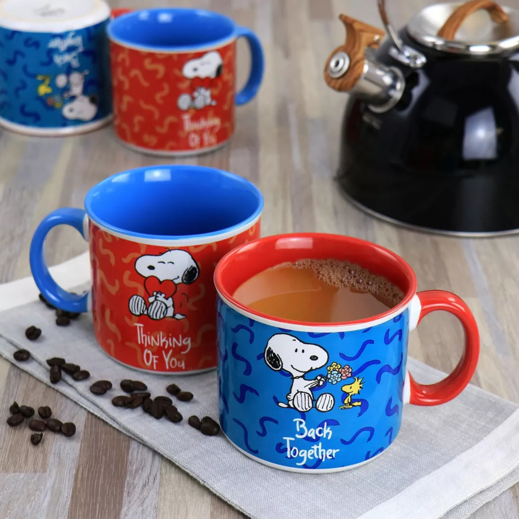 Two Snoopy-themed mugs on a wooden surface with coffee beans and a teapot.