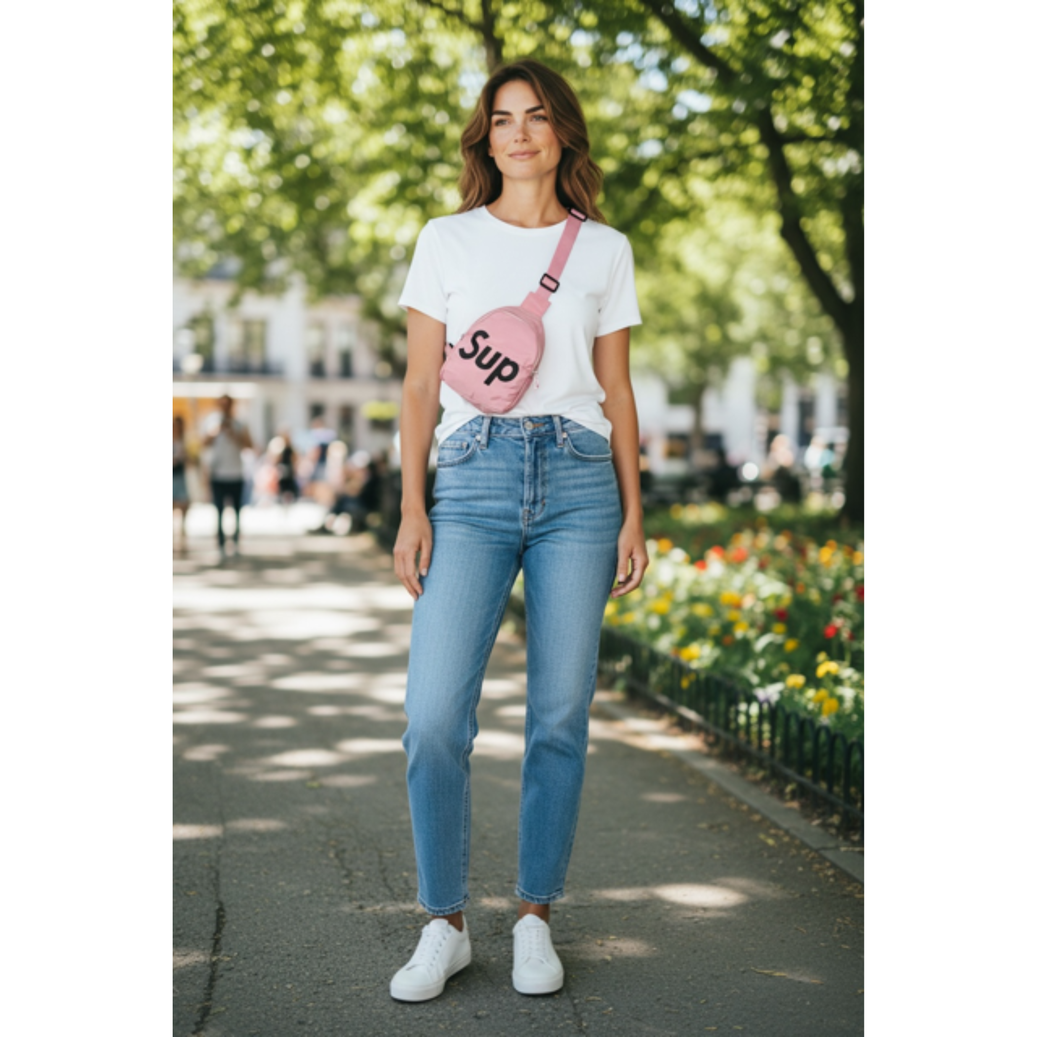 Woman wearing a white t-shirt and blue jeans, while wearing a pink sling bag with the logo 'Sup' in black letters. Woman is standing outdoors with trees and flowers in the background